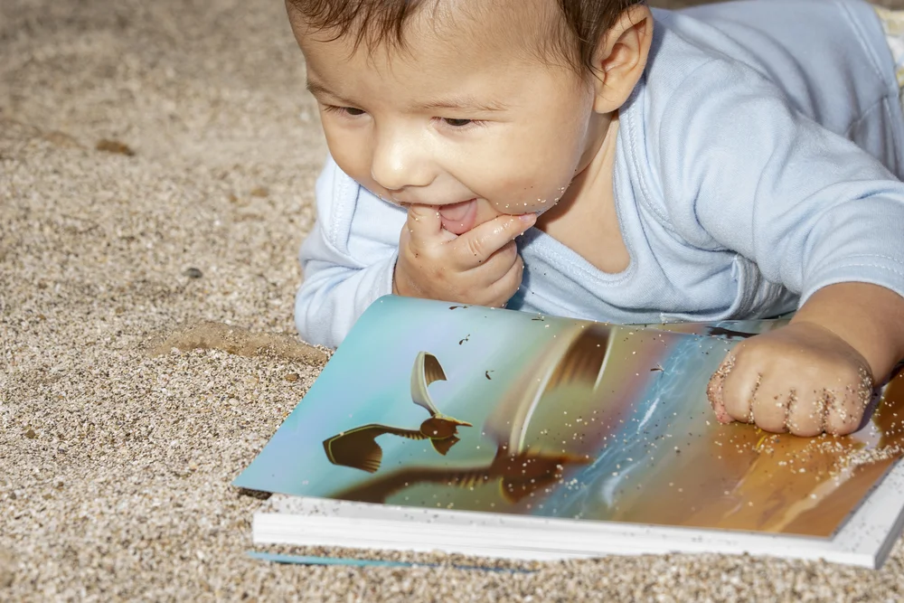 Bebé en la arena de la playa mirando sonriente el cuento Si los peces hablaran