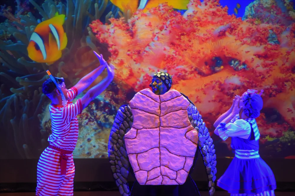 Escena del musical con la tortuga y dos bailarinas ante un arrecife de coral iluminado
