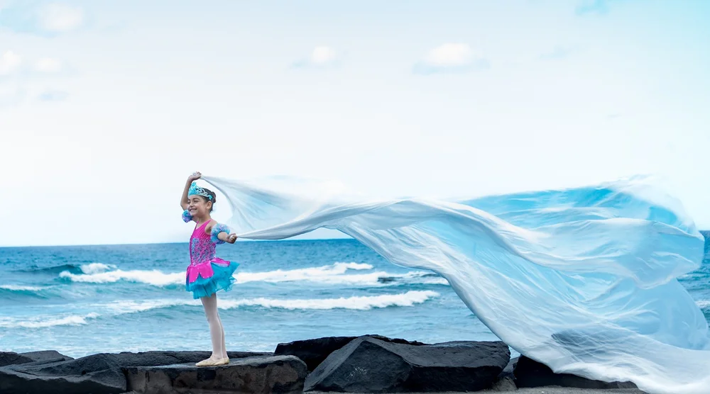 Una niña con corona y tutú sentada en un sillón frente al mar