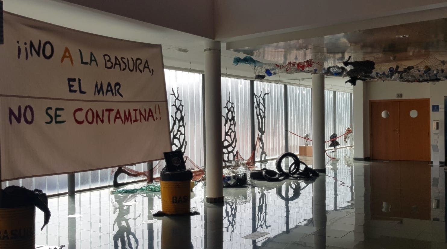 Día Mundial de los Océanos 2022_Si los pecesa hablaran_Consejería de Educación Foto del hall del Auditorio de Teror con elementos de contaminación marina acompañado de gran pancarta que está metida dentro de unos barriles de aceite que dice: ¡¡No a la basura, el mar no se contamina!! Del techo cuelga una red llena de plásticos por la que hay que pasar antes de entrar al patio de butacas, pasando primero por unas redes con nasas y gran cantidad de neumáticos. de esta manera Si los peces hablaran... inicia su labor de concienciación antes del propio espectáculo.